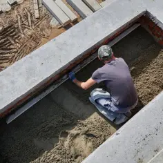 Budowlaniec wyrównuje świeży beton w wykopie. Widać ceglane ściany i betonowe elementy konstrukcji.