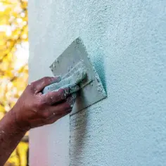 Dłoń z kielnią nakłada tynk na ścianę. W tle widać jesienne drzewa. Różne rodzaje tynków zewnętrznych dają różne efekty.