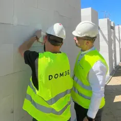 Dwóch panów w kamizelkach odblaskowych i kaskach, jeden z nich jako konstruktor budowlany sprawdza dokumenty przy ścianie z bloczków.