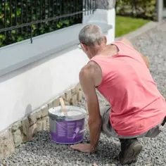 Mężczyzna w różowej koszulce maluje szarym tynkiem górną część białego, betonowego płotu. Obok stoi wiadro z farbą.