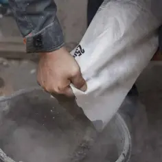 Ręce wsypują cement z worka do wiadra. Różne rodzaje cementu czekają na użycie.