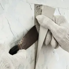 Ręce w rękawiczkach zrywają starą tapetę za pomocą noża.
