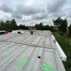 Budowa domu w toku: płyty stropowe filigran z zielonymi oznaczeniami czekają na beton. W tle drzewa i pochmurne niebo.