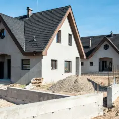 Dwa nowe domy wielorodzinne w stanie surowym, z szarymi dachami i jasnymi elewacjami, na tle błękitnego nieba.