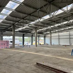 Wnętrze w budowie hali magazynowej. Betonowe słupy, stalowa konstrukcja dachu z świetlikami, ściany z płyt warstwowych. Na podłodze piasek i materiały budowlane.
