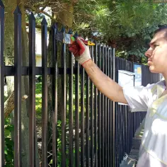 Mężczyzna w rękawicach maluje czarną farbą metalowe ogrodzenie. Przed malowaniem warto metal czymś odtłuścić, by farba lepiej się trzymała.