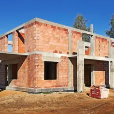 Na terenie budowy wznosi się dom z cegły ceramicznej. Betonowe stropy i słupy tworzą konstrukcję, a otwory okienne i drzwiowe czekają na wykończenie.