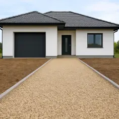 Nowy dom z białym tynkiem, czarnym dachem i bramą garażową. Szeroki podjazd do domu wysypany jasnym żwirem.