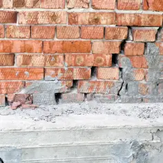Uszkodzona ceglana ściana z widocznym pęknięciem w betonie, wskazującym na podbijanie fundamentów.