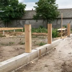 Solidny fundament pod ogrodzenie z drewnianymi słupkami, w tle budowa domu.