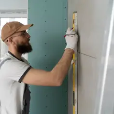 Gipsar w okularach i czapce z daszkiem mierzy ścianę poziomicą, przygotowując ją do dalszych prac.