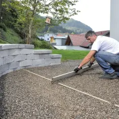 Mężczyzna wyrównuje żwir na przyszłym podjeździe z betonu, obok kamiennego murku.