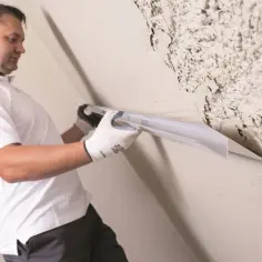 Mężczyzna w białej koszulce i rękawiczkach wykonuje gruntowanie przed malowaniem, wygładzając ścianę narzędziem.