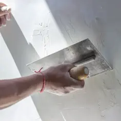Ręka z kielnią wykonuje szpachlowanie ścian, tworząc gładką powierzchnię.