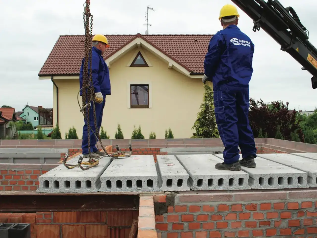 Dwóch budowlańców w żółtych kaskach montuje strop prefabrykowany na budowie domu.