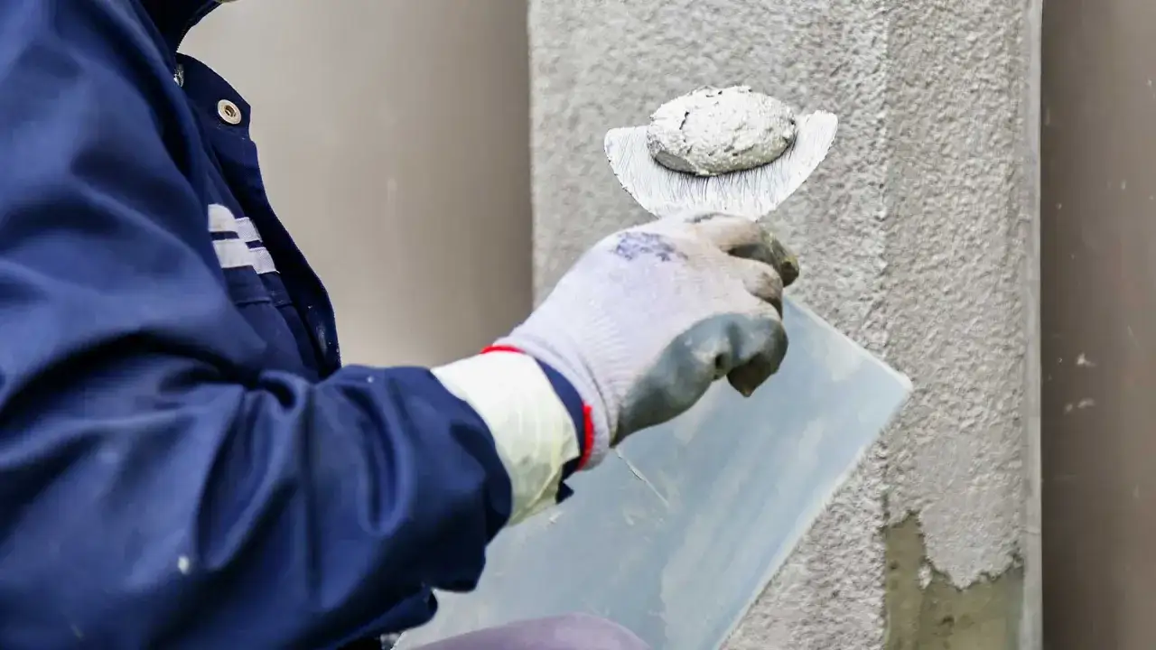 Ręka w rękawicy nakłada tynk silikonowy na ścianę za pomocą kielni.