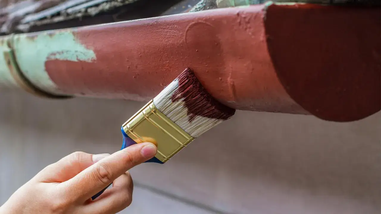 Ręka maluje pędzlem rynny aluminiowe na brązowo, odnawiając ich wygląd.