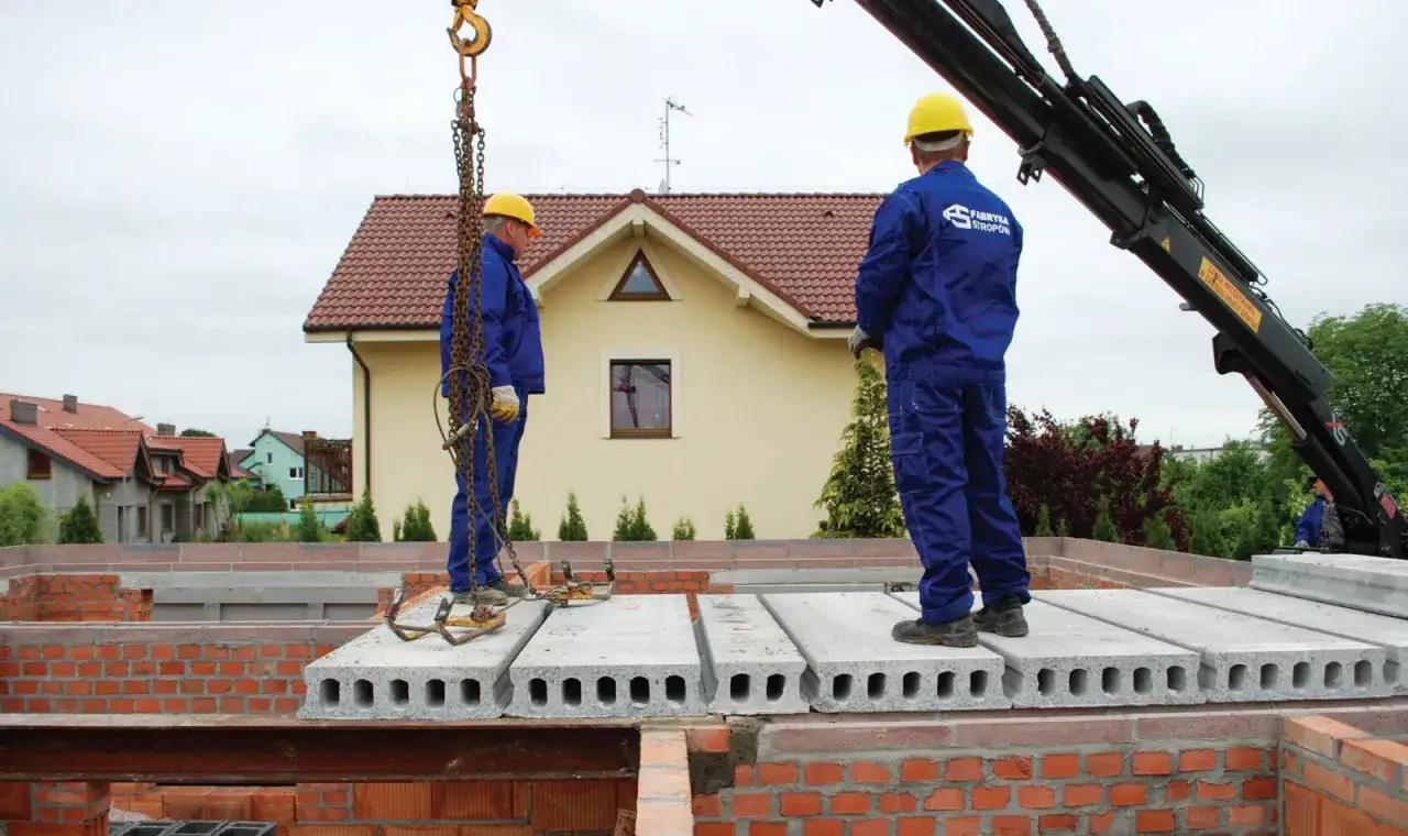 Dwóch budowlańców w żółtych kaskach i niebieskich kombinezonach montuje płyty stropowe. Dźwig podnosi kolejne elementy stropu.