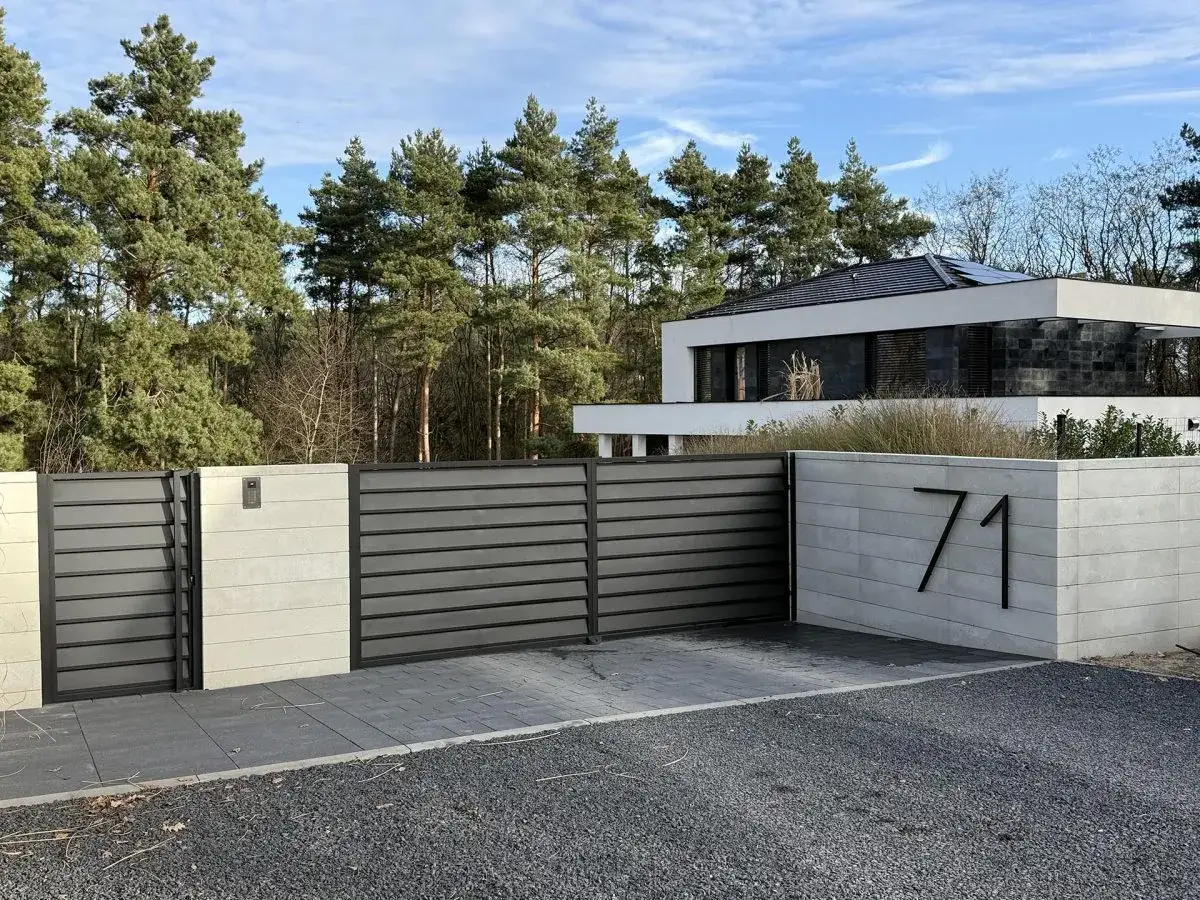 Nowoczesny dom z betonowym podjazdem, bramą i numerem 71. Otoczony sosnami.