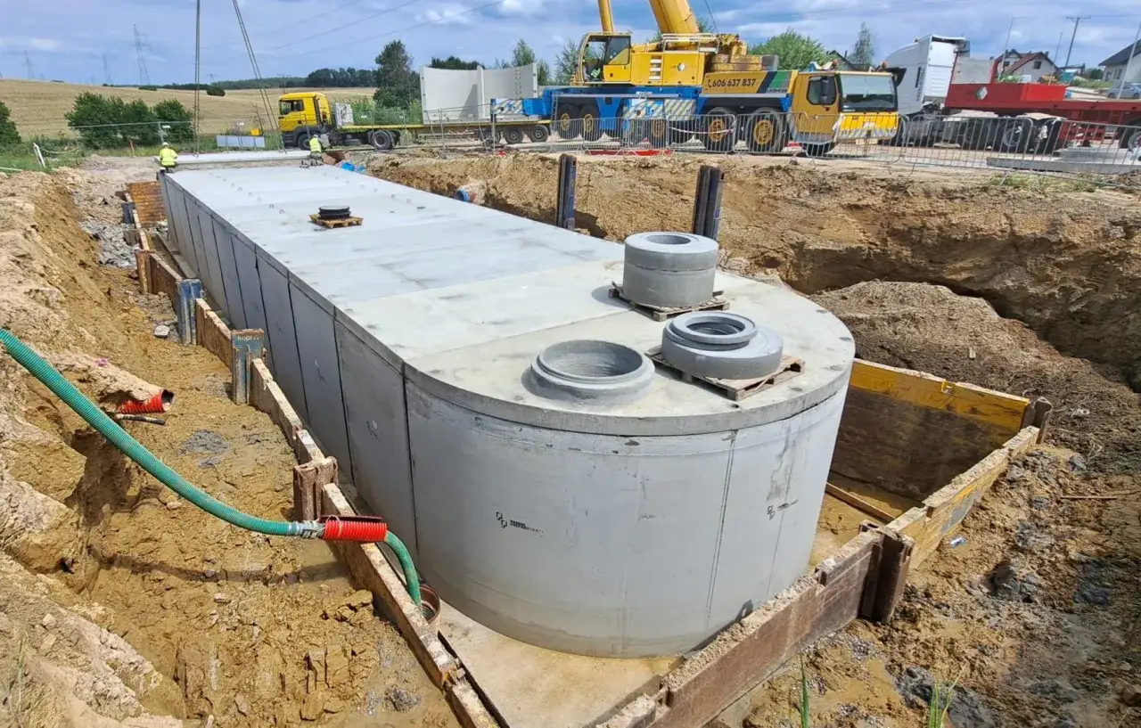 Duży betonowy zbiornik w wykopie, gotowy do montażu. Kręgi betonowe cena – kluczowy element budowy. Dźwig i koparka na placu budowy.